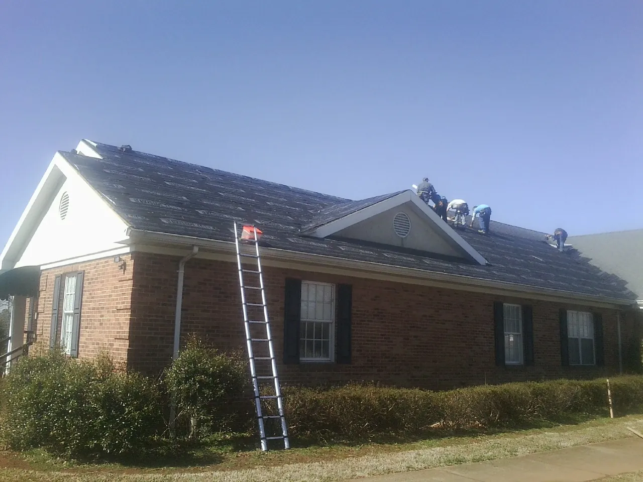 Crew tearing shingles off a brick ranch roof with a ladder set — Emergency Roof Repair work by our crew in Clearville