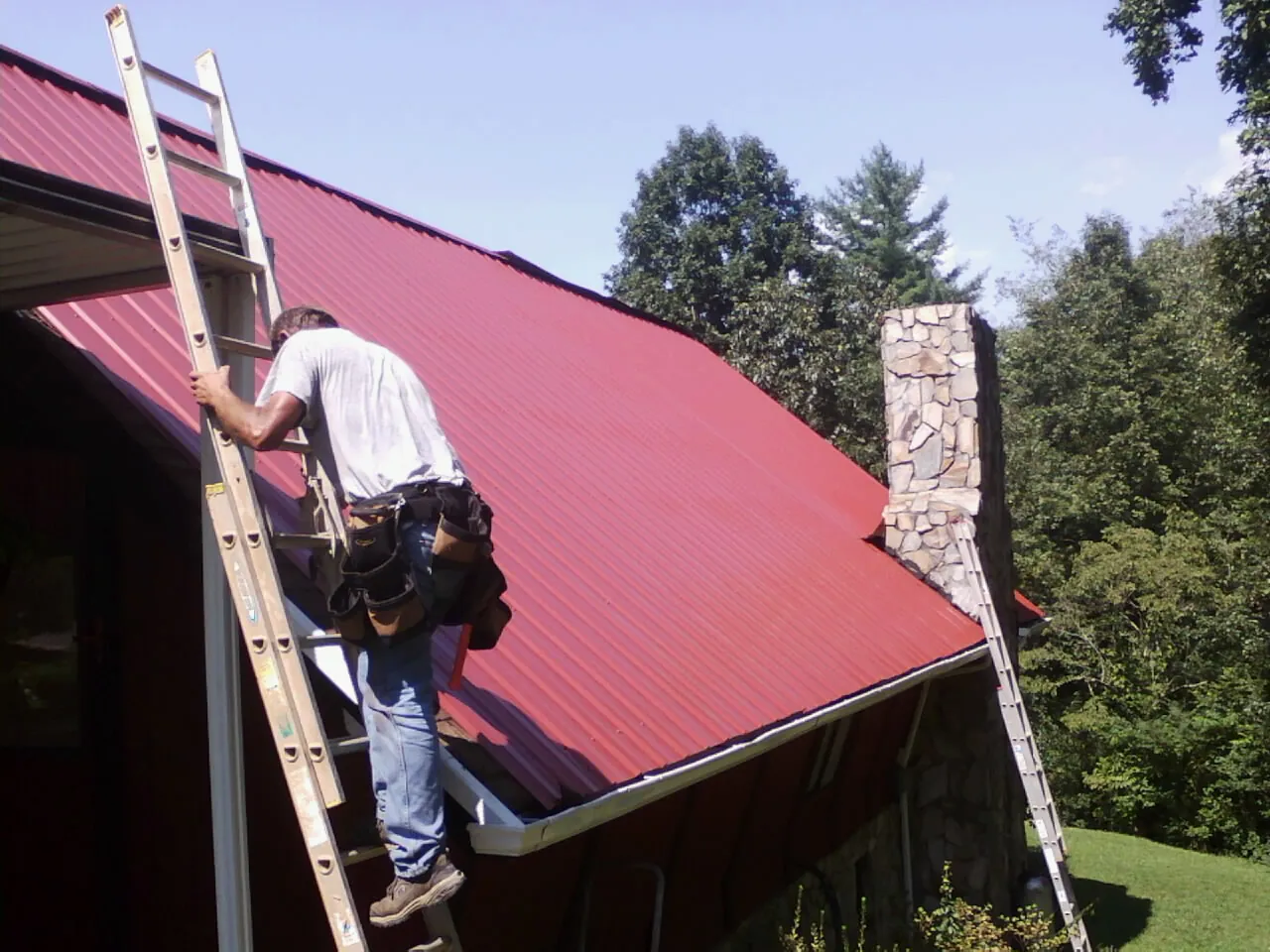 Roofer on a ladder beside a red metal roof and stone chimney — Soffit & Fascia Repair by Roofing Brothers in Clearville